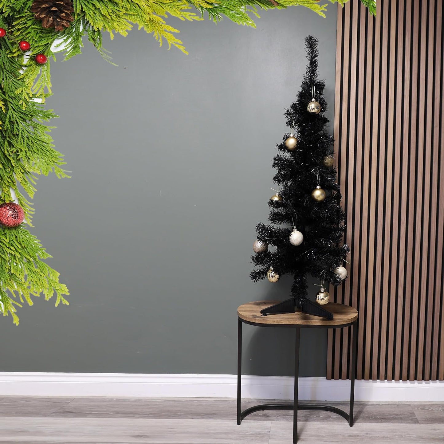 Black Christmas tree with gold and silver ornaments on a wooden table against a gray wall.