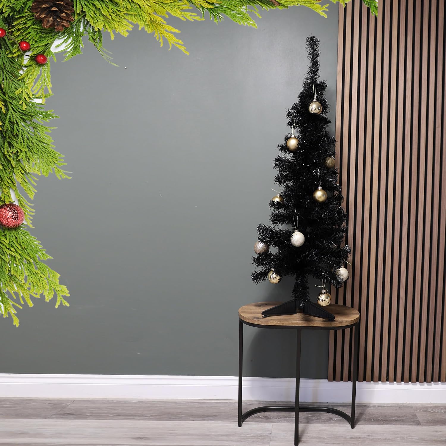 Black Christmas tree with gold and silver ornaments on a wooden table against a gray wall.