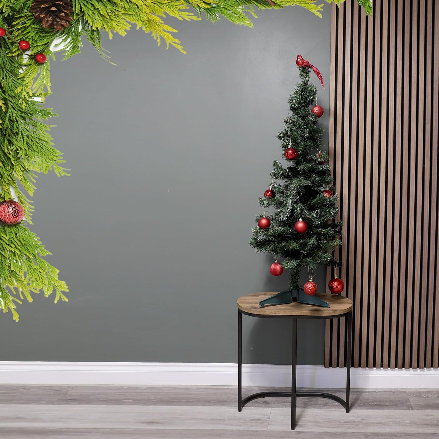 Small decorated Christmas tree on a wooden stand against a gray wall with decorative greenery.